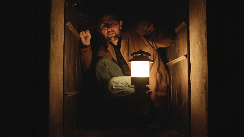 Ohm kneeling in a cramped dumbwaiter and holding a lamp in Hokum