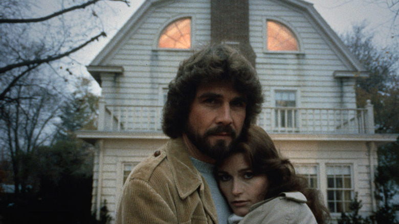 A couple embrace in front of their house while looking right into the camera.