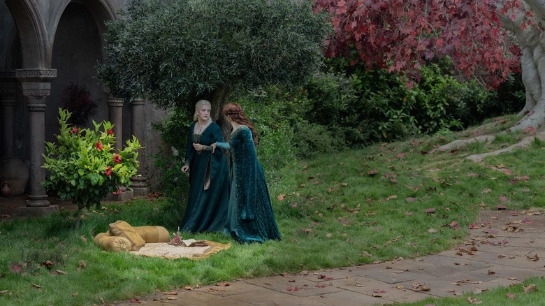 Olivia Cooke as Alicent Hightower grabbing onto the arm of Phia Saban as Helaena Targaryen while walking through a courtyard filled with trees in House of the Dragon