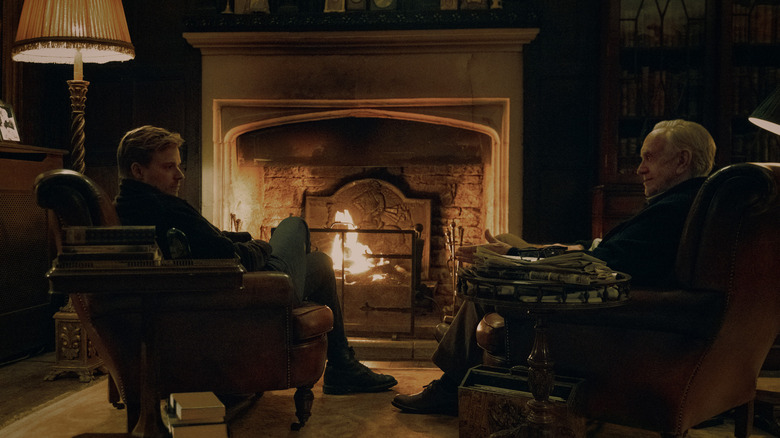 Jack Lowden as River Cartwright sitting in a lounge chair in front of a fireplace opposite Jonathan Pryce as David Cartwright in Slow Horses