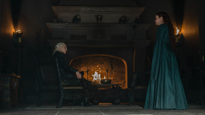 אמה ד'ארסי בתור Rhaenyra Targaryen יושבת על כיסא ליד אח מול אוליביה קוק בתור אליסנט Hightower בבית הדרקון