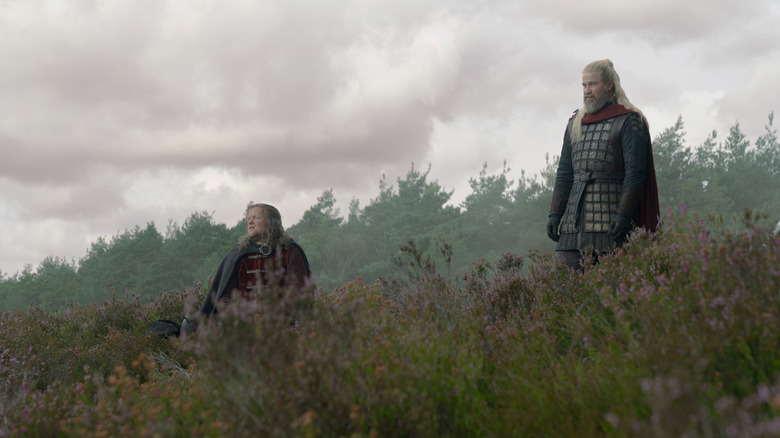Tom Bennett as Ulf sitting in an open meadow next to Kieran Bew as Hugh wearing armor in House of the Dragon