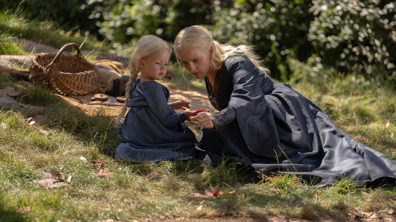 Phia Saban as Helaena Targaryen seated on a grassy hill next to Ziggy Barker as Jaehaera Targaryen enjoying a picnic in House of the Dragon