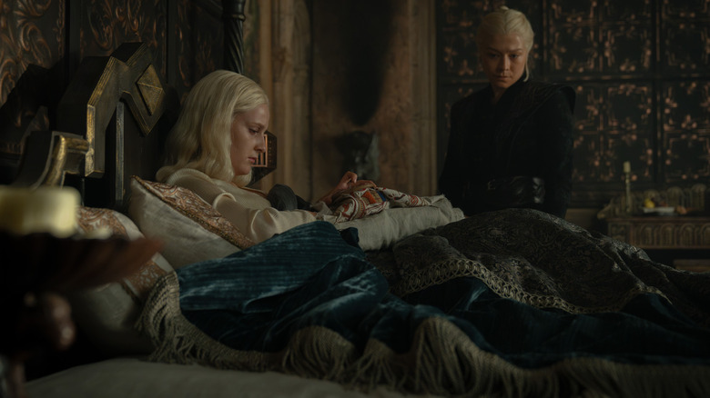 Emma D'Arcy as Rhaenyra Targaryen sitting beside Phia Saban as Helaena Targaryen knitting a tapestry in bed in House of the Dragon