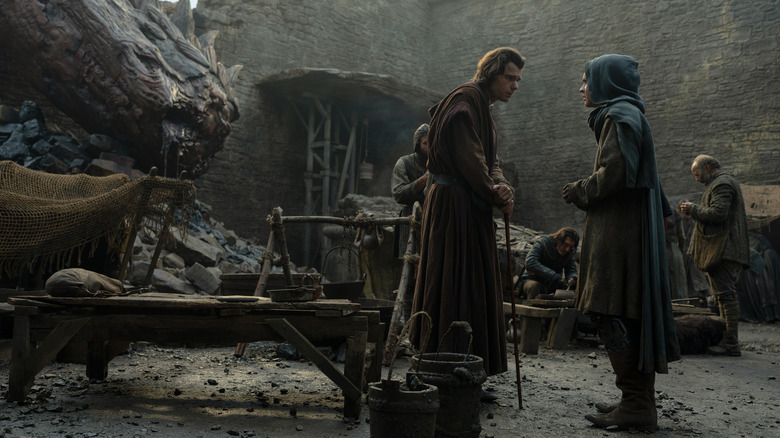 Matthew Needham as Larys Strong leans on his cane opposite Tom Glynn-Carney as Aegon Targaryen in a hood while the remains of a dead dragon lay behind them in House of the Dragon