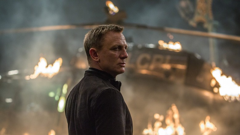 Daniel Craig as James Bond standing in front of flaming wreckage in Spectre