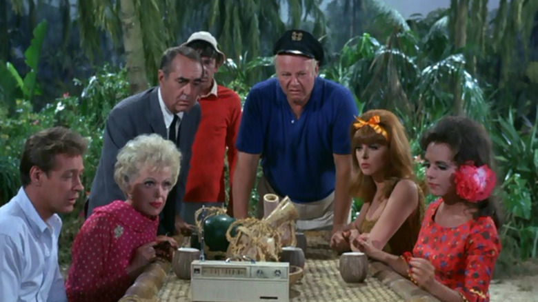 The cast of Gilligan's Island gathered around a table on the island, looking puzzled