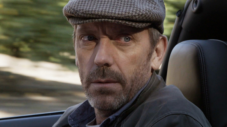 Hugh Laurie as House sits in a car wearing a flat cap in House