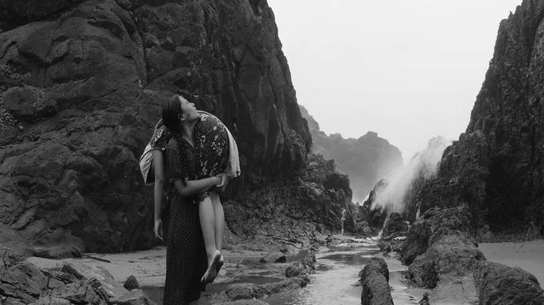 A woman carrying someone over her shoulder through large rocks and a river in From What Is Before