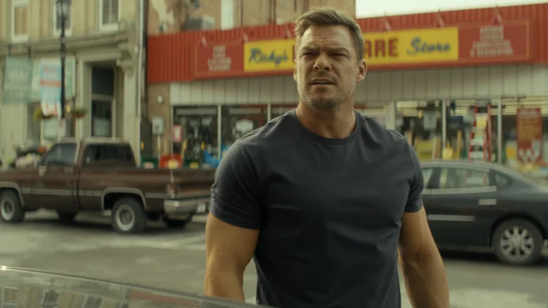 Alan Ritchson as Jack Reacher stands beside a car in a street in Reacher