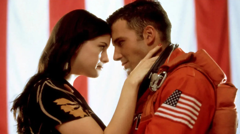 Grace (Liv Tyler) and A.J. (Ben Affleck), wearing an orange astronaut suit, lean in to touch foreheads while standing in front of a large American flag backdrop in Armageddon