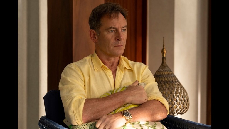 Timothy Ratliff (Jason Isaacs) sits clutching a pillow, his anxious expression clear, wearing a bright yellow shirt in The White Lotus