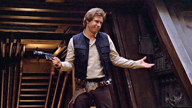 Han Solo (Harrison Ford) shrugs and has a cocky grin as he shrugs while holding his blaster and standing in a spaceship corridor in Star Wars: Episode IV – A New Hope