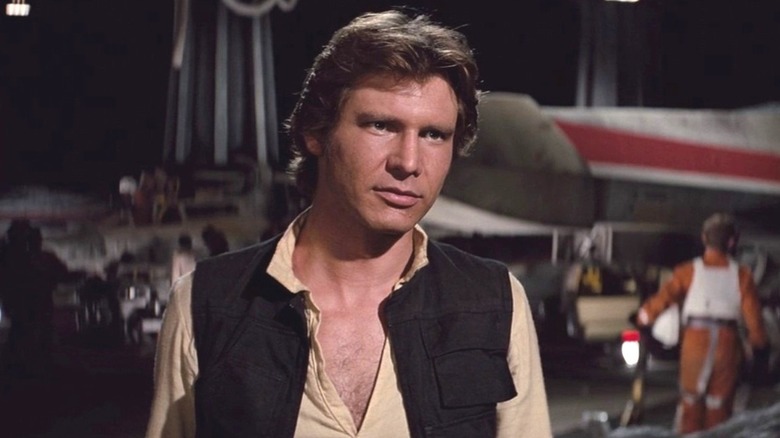 Han Solo (Harrison Ford) stands in front of a Rebel Alliance hangar wearing a skeptical expression in Star Wars: Episode IV – A New Hope