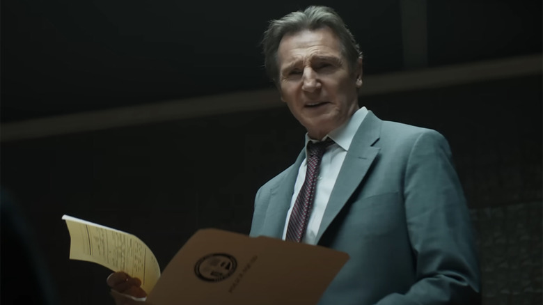 Liam Neeson as Frank Drebin, smirking while holding a folder in an interrogation room in The Naked Gun (2025)