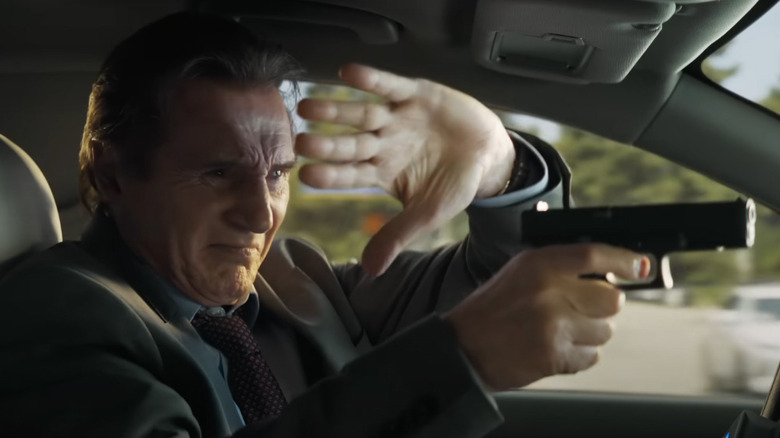 Liam Neeson as Frank Drebin shoots a gun from the driver's seat of a car while driving in The Naked Gun (2025)