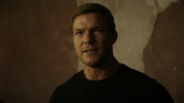 అలాన్ రిచ్సన్ యొక్క జాక్ రీచర్ రీచర్‌లోని సాదా గోడకు ఆనుకుని నిల్చున్నప్పుడు క్లోజప్‌లో కనిపించాడు