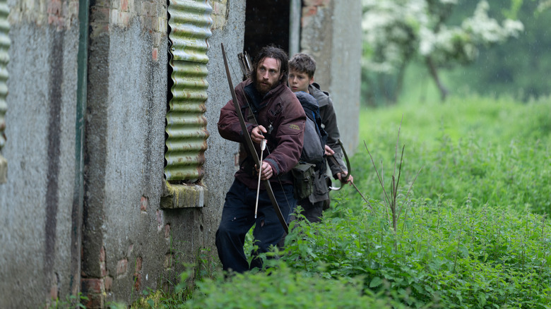Aaron Taylor-Johnson as Jamie and Alfie Williams as Spike holding bows and arrows and walking beside an abandoned house in 28 Years Later