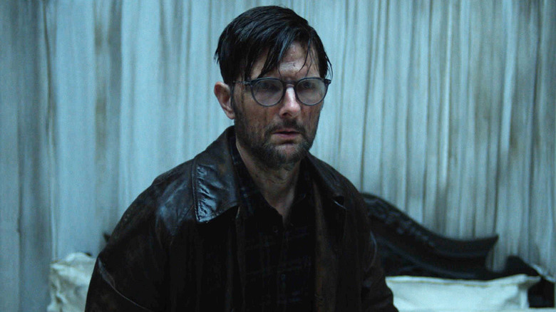Adam Scott's Ohm Bauman stands in a room in front of a dirty curtain in Hokum