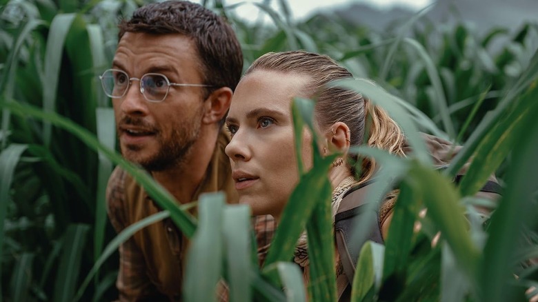 Jonathan Bailey as Dr. Henry Loomis and Scarlett Johansson as Zora Bennett crouching in tall grass in Jurassic World Rebirth