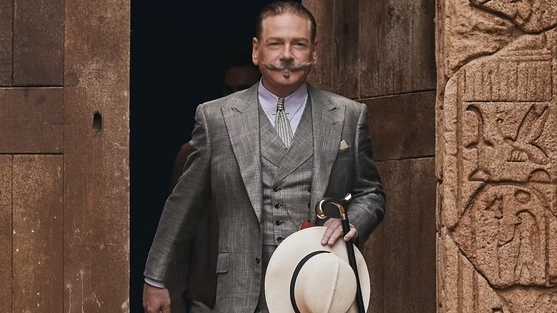 Hercule Poirot holding his hat and cane which leave a building covered with hieroglyphs in death on the Nile