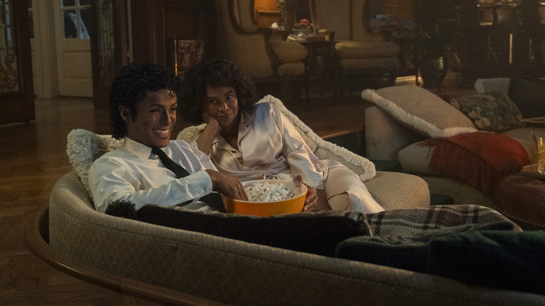 Jaafar Jackson as Michael Jackson eating popcorn and smiling while sitting on a couch next to Nia Long as Katherine Jackson in Michael