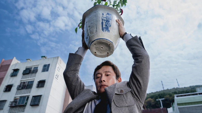 Lee Byung-hun as Yoo Man-su lifting a potted plant over his head in No Other Choice
