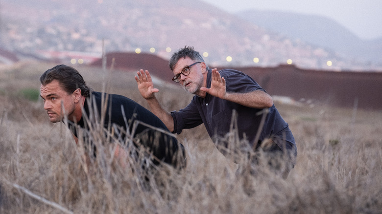 Director Paul Thomas Anderson crouching in a field with Leonardo DiCaprio as Bob Ferguson on the set of One Battle After Another