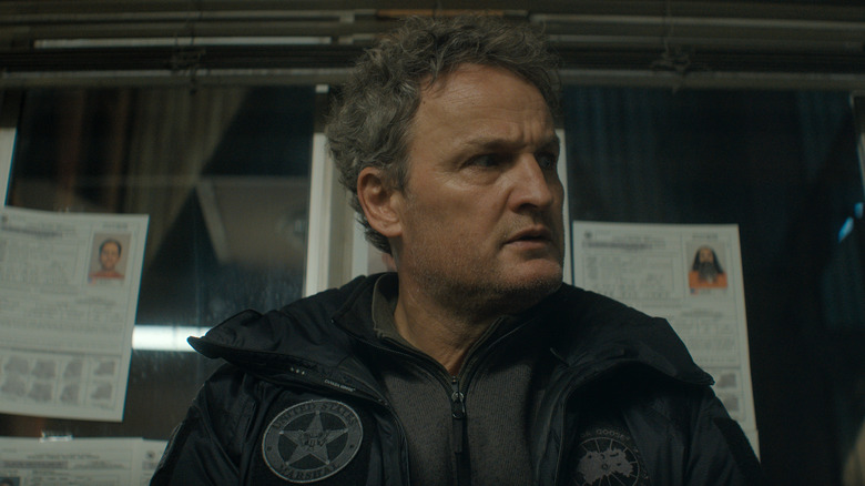 Jason Clarke as Frank Remnick seated in front of a window with pictures of mugshots in The Last Frontier