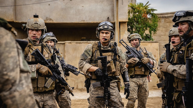 Charles Melton's Jake stands in the center of the frame, holding a gun and surrounded by fellow soldiers, in Warfare