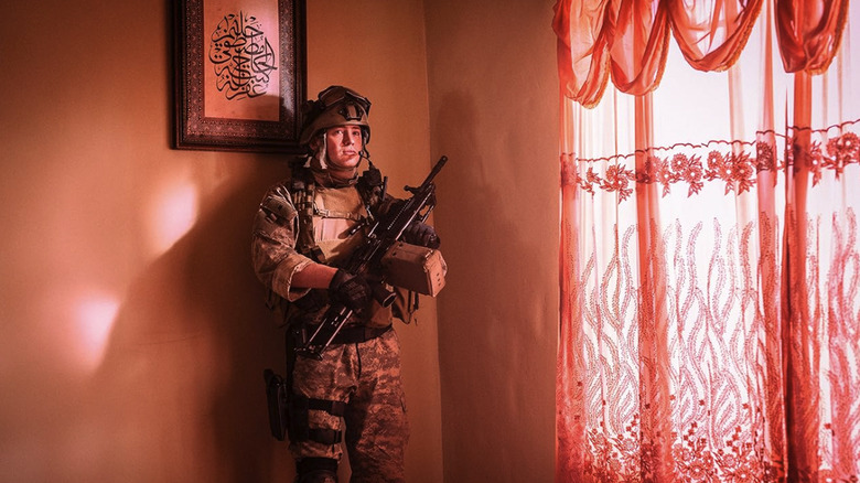 Kit Connor's Tommy stands in the corner of a room, bathed in red light and holding a gun, in Warfare