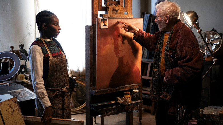 Michaela Coel as Lori Butler standing in a cluttered room next to Ian McKellen as Julian Sklar gesturing at a painting in The Christophers