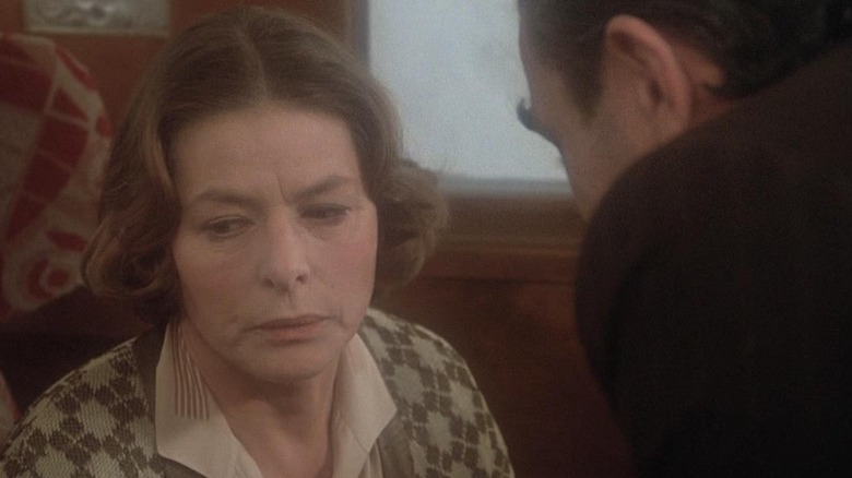 Greta Ohlsson sits in a train car, looking off to her right, while a man bends down and talks to her in Murder On the Orient Express