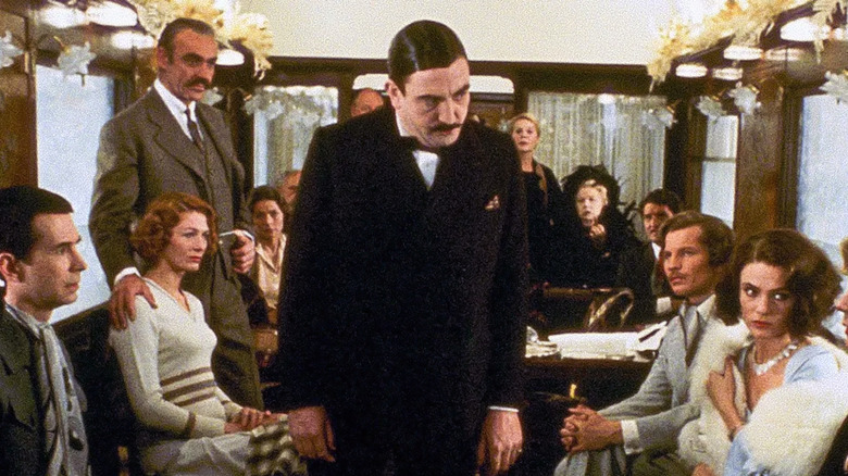 A group of well -dressed people is sitting in an opulent train car and looks at Hercule Poirot, standing in the middle and addressing them all in the murder on the East Express