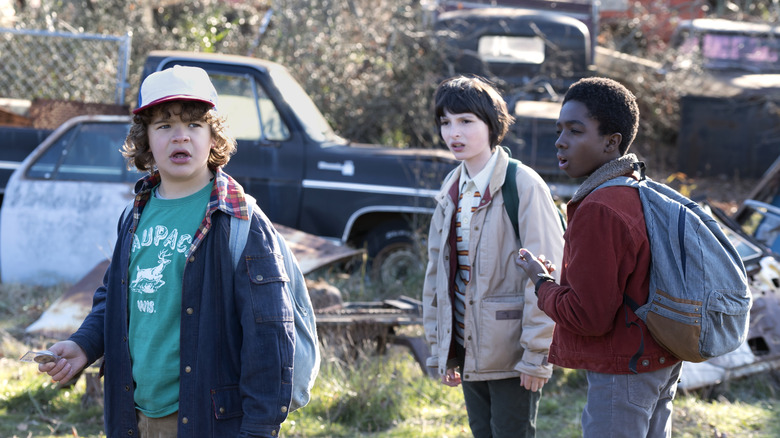 Dustin, Mike, and Lucas wandering a scrap yard in Stranger Things season 1