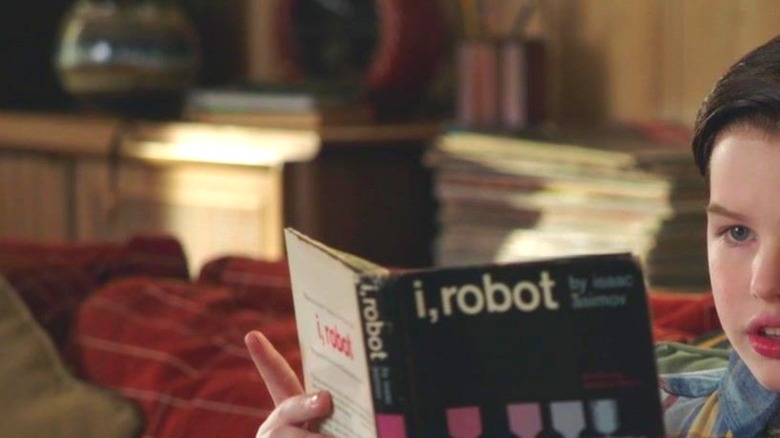 Iain Armitage as Sheldon Cooper reading a library copy of I, Robot by Isaac Asimov on his couch on the series Young Sheldon
