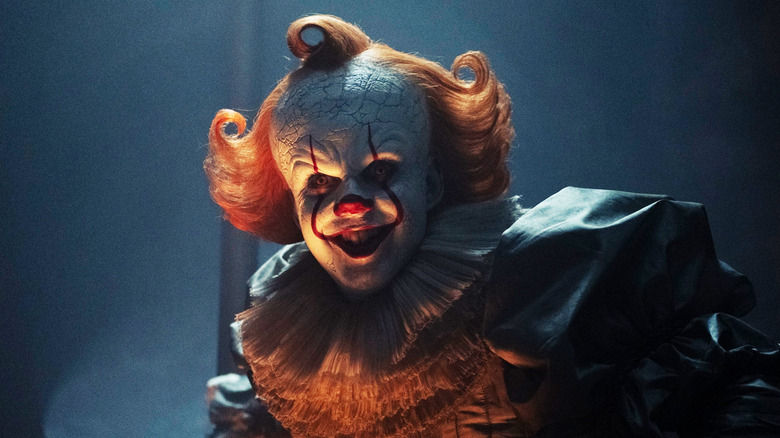 Bill Skarsgård's Pennywise grins as he crouches in a dark room in It Welcome to Derry