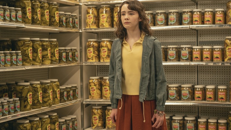 Clara Stack as Lilly Bainbridge standing in a grocery store surrounded by pickle jars in It: Welcome to Derry