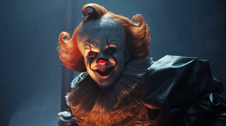 Bill Skarsgård's Pennywise grins as he crouches in a dark room in It: Welcome to Derry
