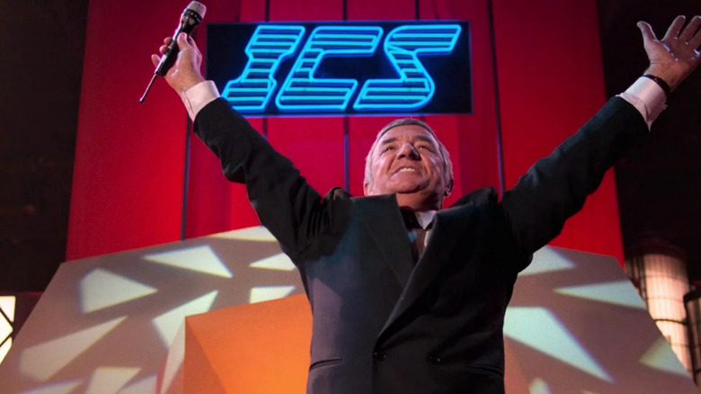 Richard Dawson as Damon Killian in front of light up letters that read ICS in The Running Man