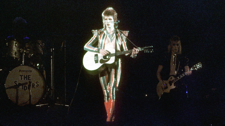 man in a striped blazer with no shirt holding a guitar in front of a microphone
