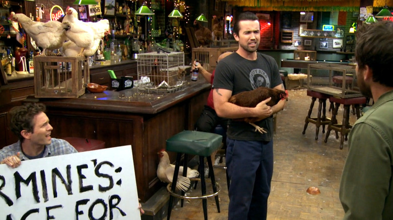 Always Sunny, Dennis holding a sign, Mac holding a rooster, a bunch of loose chickens on the bar, Charlie's looking on in confusion