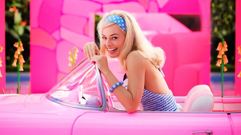 margot robbie as barbie smiling at the camera in a pink convertible in front of a pink stone wall