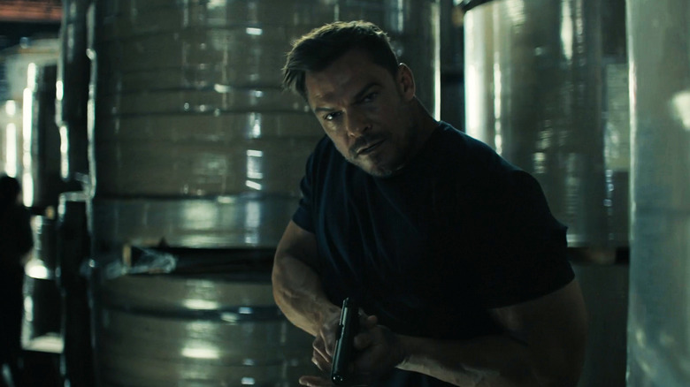 Alan Ritchson's Jack Reacher holds a gun as he walks through a warehouse in Reacher