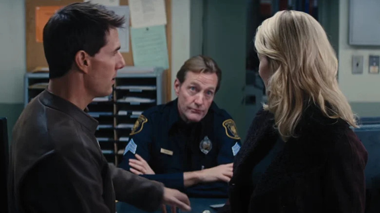 Lee Child's desk sargeant is seen sitting at a desk between Tom Cruise's Jack Reacher and Rosamund Pike's Helen Rodin in Jack Reacher.webp