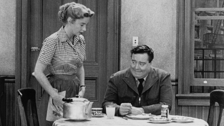 Ralph at a dinner table next to a standing Alice in The Honeymooners