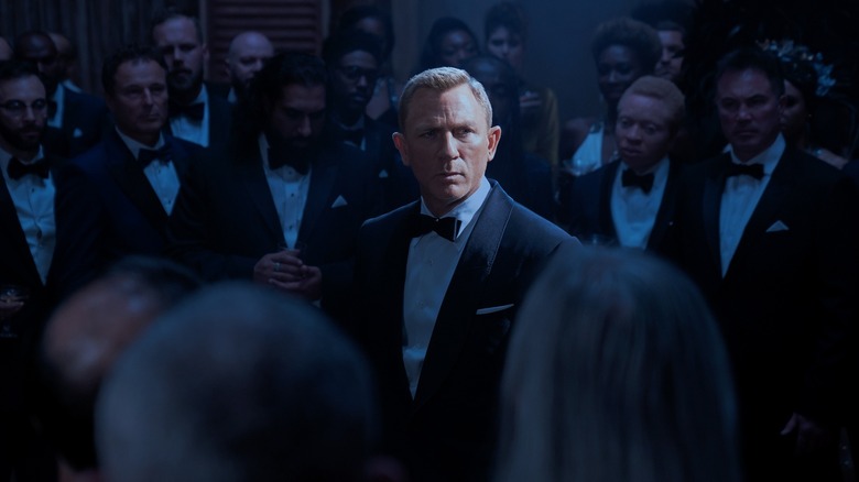 Daniel Craig as James Bond standing in the middle of a crowd of onlookers all dressed in tuxedos in Spectre