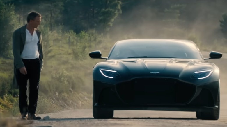 Daniel Craig as James Bond walking next to a 2020 Aston Martin DBS Superleggera on a dirt road in No Time to Die