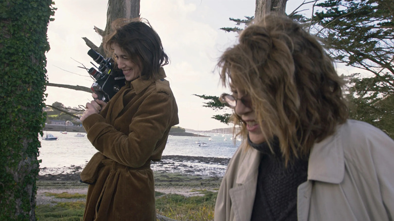 two women in the woods walking next to one another, the woman in the background is holding a super 8 video camera