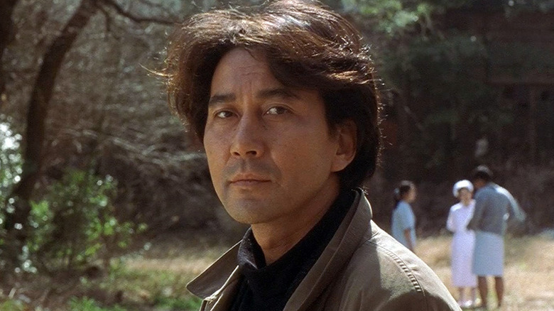 Kenichi Takabe (Kōji Yakusho) stands outside with hospital staff in the background in Cure